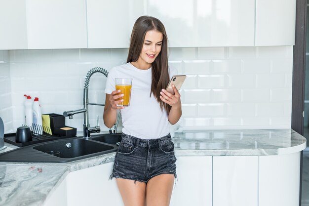 Mujer sonriente mirando el teléfono móvil y sosteniendo un vaso de jugo de naranja en una cocina
