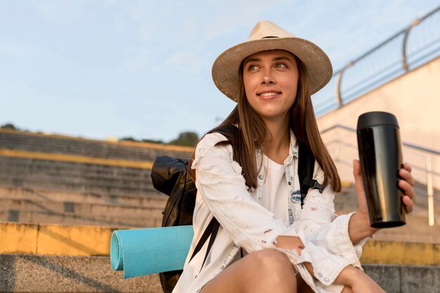 Mujer sonriente con mochila y sombrero sosteniendo termo mientras viaja