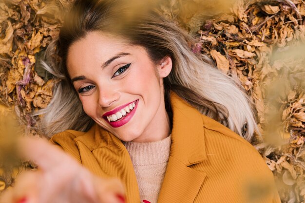 Mujer sonriente moderna que mira la cámara que miente en las hojas secas durante la estación del otoño