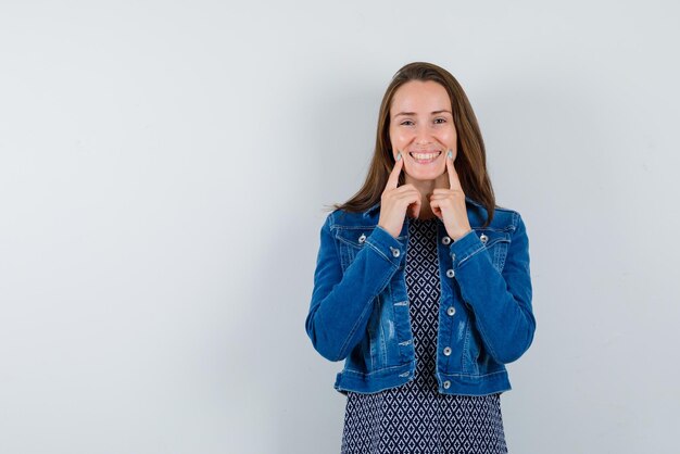 La mujer sonriente muestra su sonrisa con los dedos índices sobre fondo blanco.