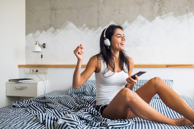 Mujer sonriente muy sexy sentada en la cama por la mañana, escuchando música en auriculares, divirtiéndose