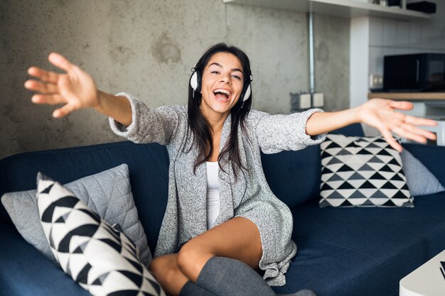 Mujer sonriente muy sexy en traje casual sentado en la sala de estar escuchando música en auriculares, divirtiéndose en casa