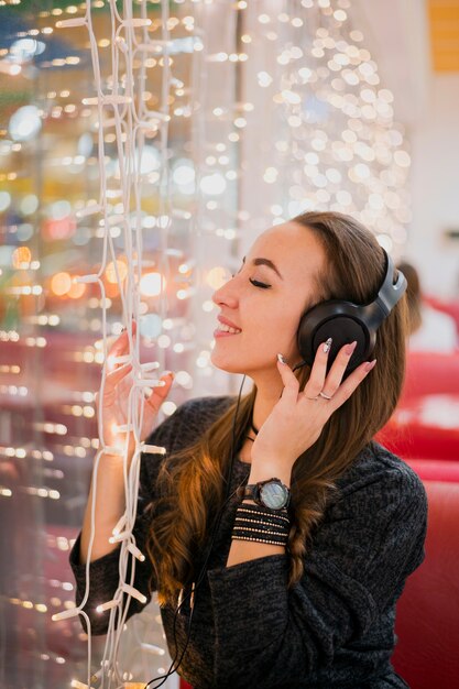 Mujer sonriente con los ojos cerrados con auriculares en la cabeza tocando las luces de navidad