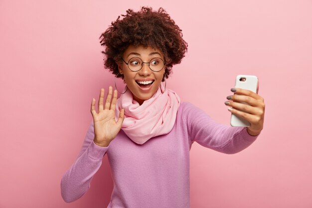 Mujer sonriente optimista dice hola graba mensaje de video, llama a través de la aplicación de redes sociales en el celular, sostiene el dispositivo en el frente, saluda con la palma, vestida con un atuendo elegante