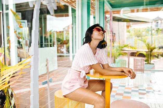 Mujer sonriente en pantalones cortos sentado en la cafetería al aire libre. Mujer soñadora en gafas de sol rosas disfrutando del fin de semana de verano.