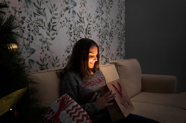 Mujer sonriente con el paquete del regalo en el settee cerca del árbol de navidad