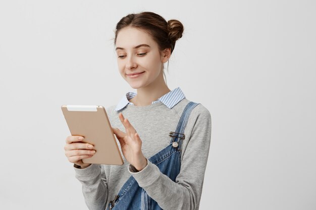 Mujer sonriente con el pelo atado en bollos dobles mirando la pantalla del dispositivo moderno. Encantadora chica cutie escribiendo mensaje a su novio usando tableta. Concepto de relaciones