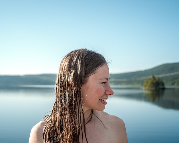 Mujer sonriente con el pelo mojado rodeado por el lago y colinas bajo la luz del sol