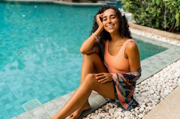 Mujer sonriente con pelos ondulados relajándose cerca de la piscina Traje elegante tropical Flor de Plumeria en los pelos
