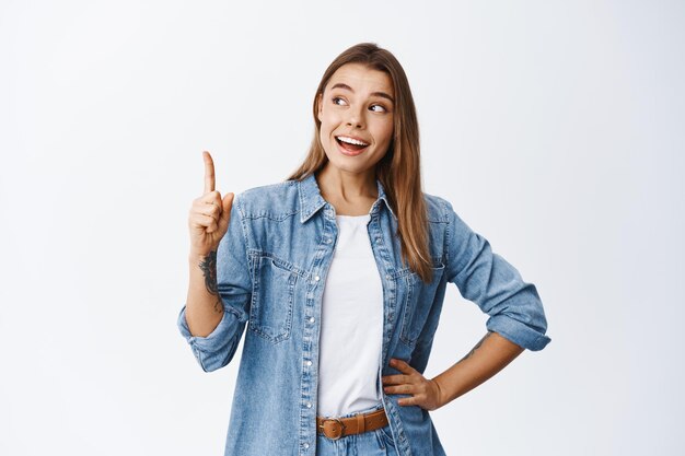 Mujer sonriente pensativa con cabello rubio, levantando el dedo y apuntando hacia arriba, teniendo un buen punto o idea, mostrando publicidad, de pie en blanco