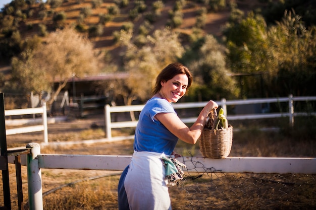 Mujer sonriente de pie cerca del rancho con canasta de vegetales cosechados