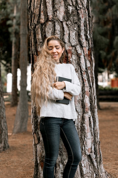 Mujer sonriente de pie junto a un árbol mientras sostiene un libro