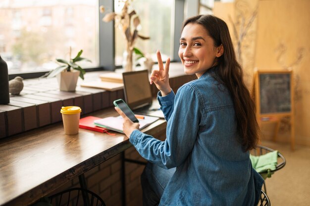 Mujer sonriente posando con smartphone en el café mientras espera a su amiga