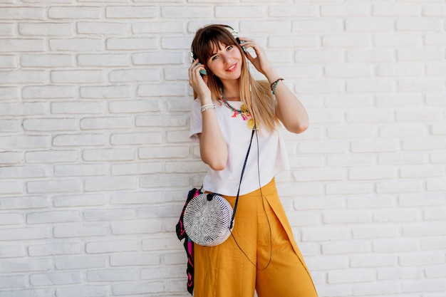 Mujer sonriente posando sobre pared de ladrillo blanco con auriculares y mostrando signos.