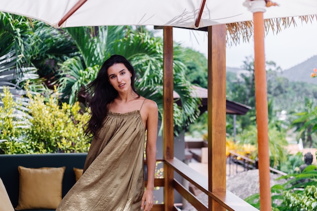 Mujer sonriente positiva bastante europea en vestido de vuelo de verano a la luz natural del día en la terraza de la villa disfrutando de hermosas vacaciones, sofá al aire libre con almohadas en tropical