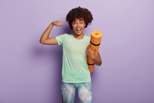 Mujer sonriente positiva con cabello afro, sostiene estera de yoga