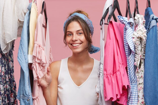 Mujer sonriente positiva con camiseta blanca y bufanda, mirando a través del riel de la ropa mientras está de pie en su probador, contenta de tener mucha ropa nueva de moda. Concepto de moda y personas