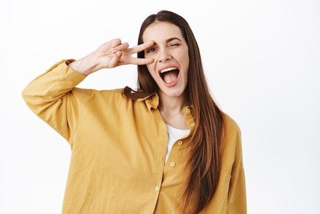 Mujer sonriente positiva que muestra un gesto de vsign kawaii y guiña la boca abierta feliz permaneciendo en el lado positivo de pie optimista contra el fondo blanco