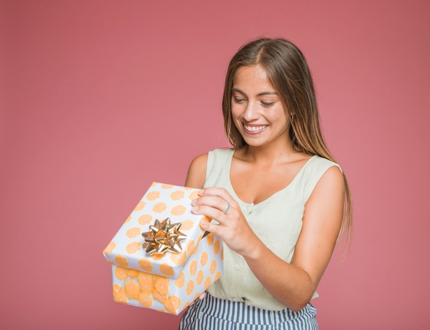 Mujer sonriente que abre la caja de regalo floral con el arco de oro contra el telón de fondo de color