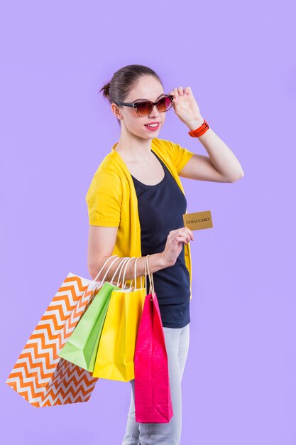 Mujer sonriente que lleva el bolso de compras colorido con sostener la tarjeta del oro en el papel pintado púrpura