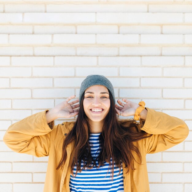 Mujer sonriente que lleva el sombrero hecho punto y la chaqueta que se colocan delante de la pared de ladrillo