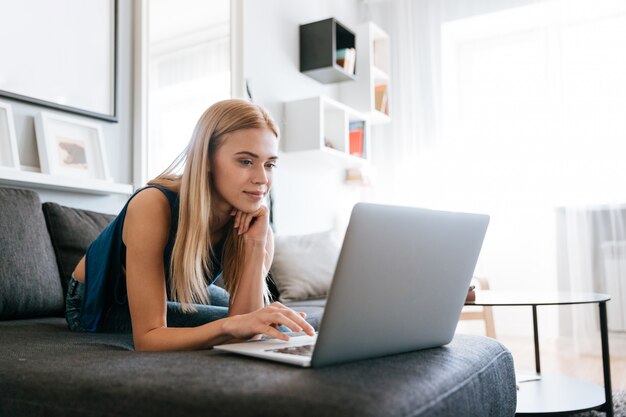 Mujer sonriente que miente en el sofá y que usa la computadora portátil en casa