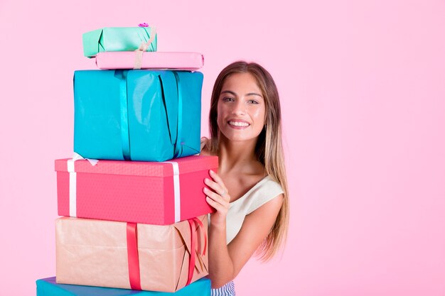 Mujer sonriente que mira a escondidas de la pila de cajas de regalo coloridas contra fondo rosado