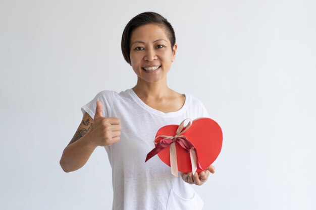 Mujer sonriente que muestra la caja y el pulgar en forma de corazón de regalo para arriba