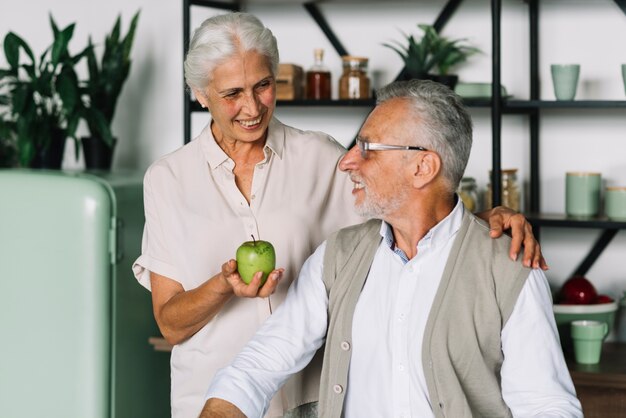 Mujer sonriente que muestra la manzana verde a su marido que se coloca en la cocina