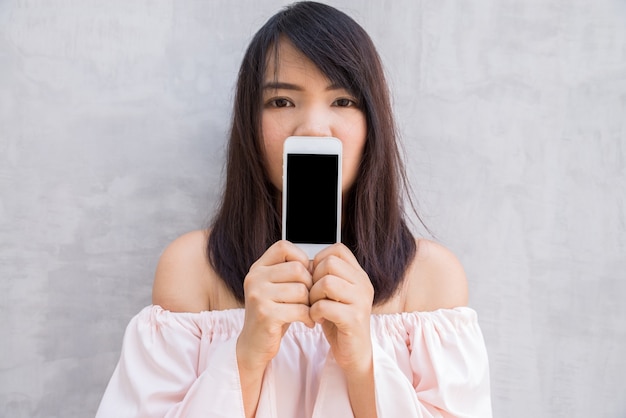 Foto gratuita mujer sonriente que muestra una pantalla en blanco del smartphone que se coloca en el muro de cemento.