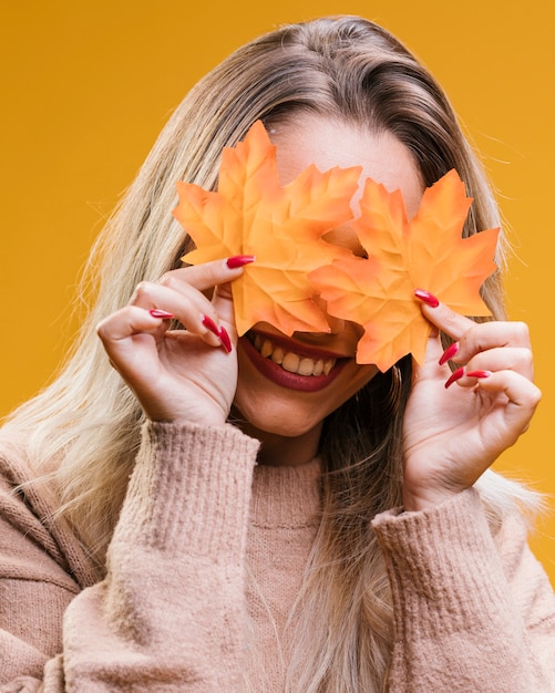 Mujer sonriente que oculta sus ojos con las hojas de arce contra fondo amarillo