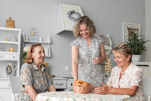 Foto gratuita mujer sonriente que pone la cesta de mimbre de croissant en la mesa frente a una mujer madura y una mujer mayor