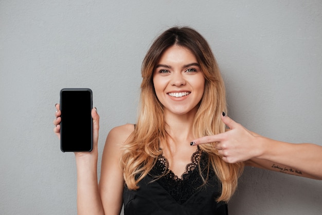 Mujer sonriente que señala el dedo en el teléfono móvil de la pantalla en blanco