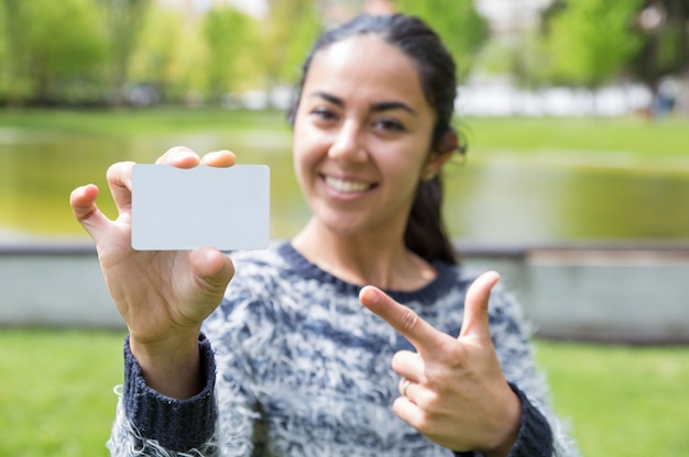 Mujer sonriente que señala en la tarjeta de visita en blanco en parque de la ciudad