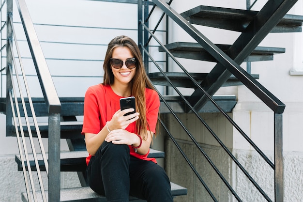 Mujer sonriente que se sienta en la escalera usando el teléfono móvil