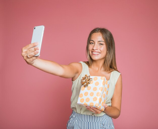 Mujer sonriente que sostiene la caja de regalo que toma el selfie del teléfono móvil contra fondo rosado