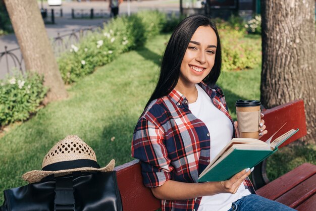 Mujer sonriente que sostiene el libro y la taza de café disponible mientras que se sienta en banco en el parque