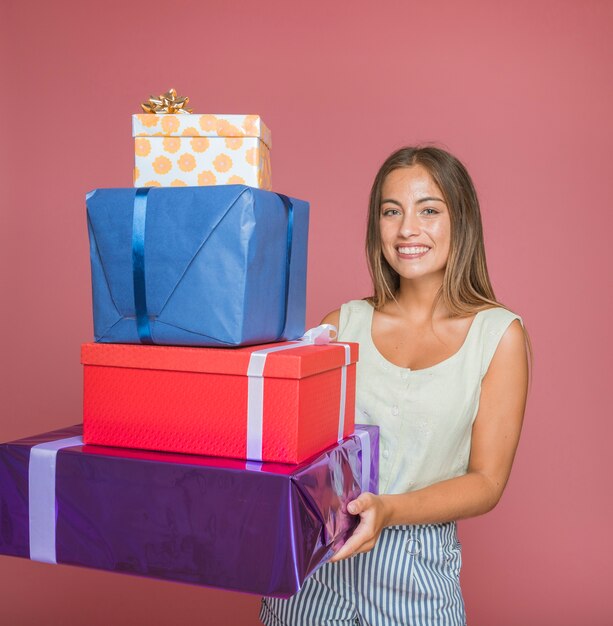 Mujer sonriente que sostiene la pila de cajas de regalo coloridas contra el contexto rosado