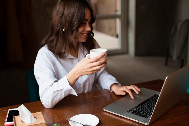 Mujer sonriente que sostiene la taza de café y que trabaja en la computadora portátil