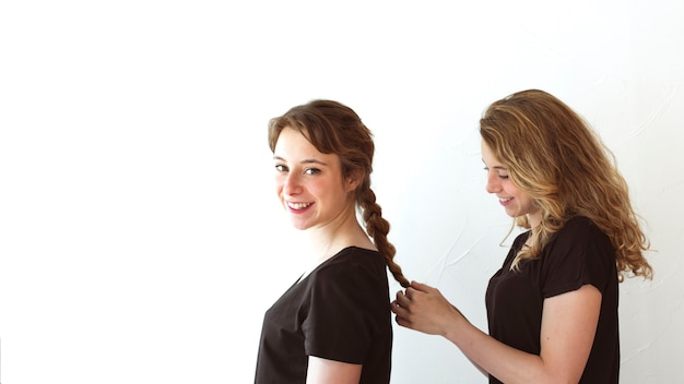 Mujer sonriente que trenza el pelo de su hermana aislada sobre el contexto blanco