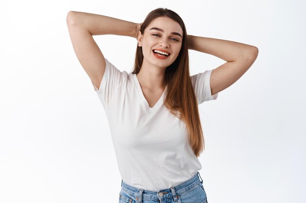 Mujer sonriente relajada que tiene tiempo libre, mantenga las manos detrás de la cabeza en reposo, pose relajante y riendo, disfrute de un fin de semana de ocio, de pie sobre fondo blanco.