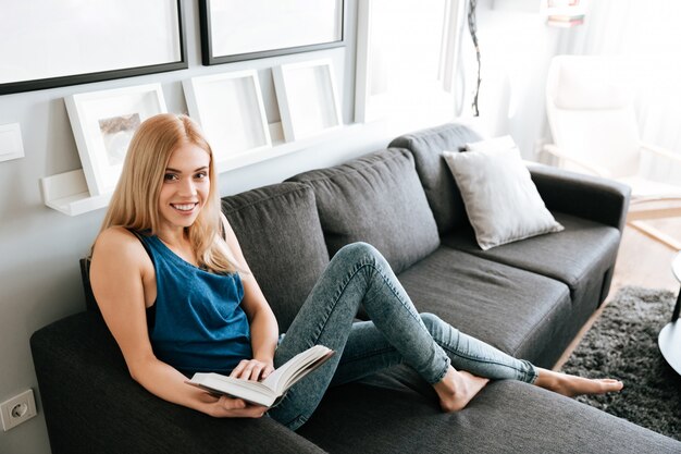 Mujer sonriente relajante y libro de lectura en el sofá en casa