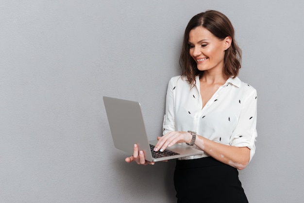Mujer sonriente en ropa de negocios posando cerca de la pared y usando la computadora portátil en gris