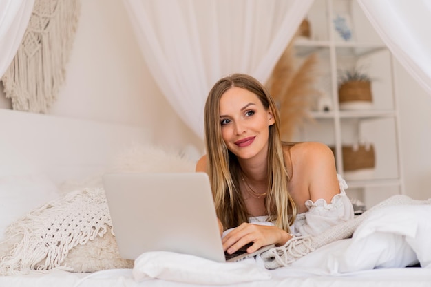 Mujer sonriente rubia acostada en una cama acogedora y usando la computadora portátil para compras en línea.
