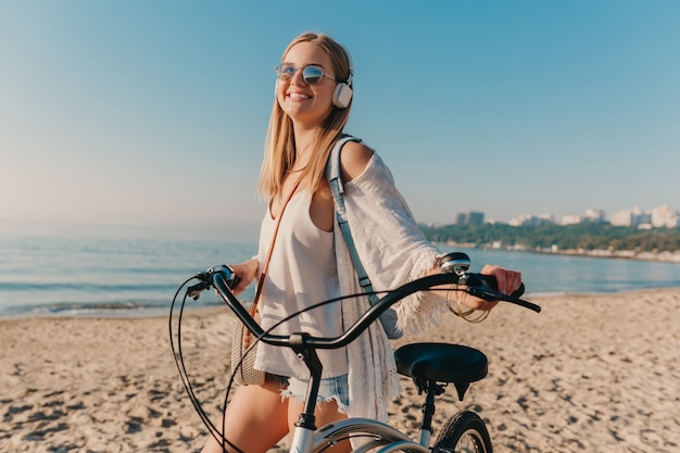 Mujer sonriente rubia atractiva joven que camina en la playa con la bicicleta en auriculares escuchando música