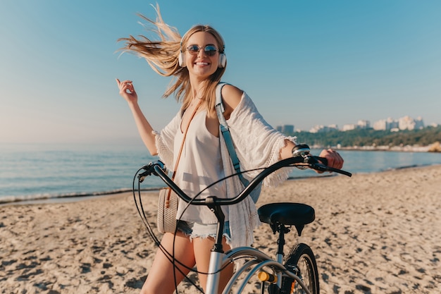 Mujer sonriente rubia atractiva joven que camina en la playa con la bicicleta en auriculares escuchando música