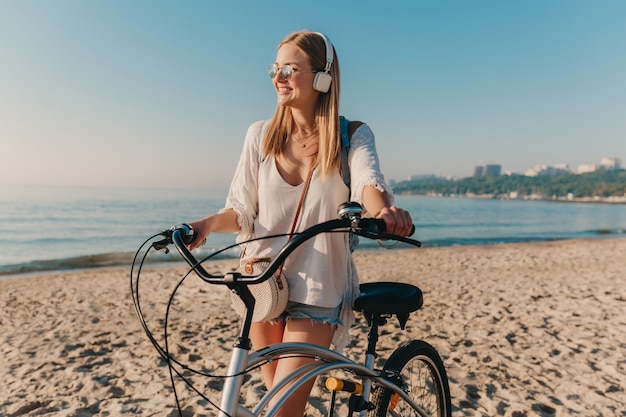 Mujer sonriente rubia atractiva joven que camina en la playa con la bicicleta en los auriculares