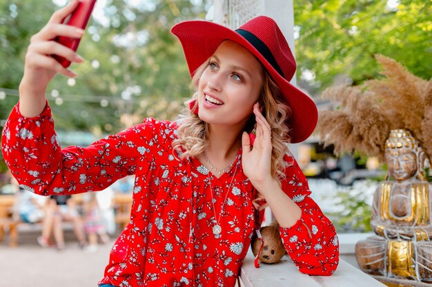 Mujer sonriente rubia elegante atractiva en traje de la manera del verano de la blusa y del sombrero rojo de la paja que toma la foto del selfie en café del teléfono inteligente de la cámara