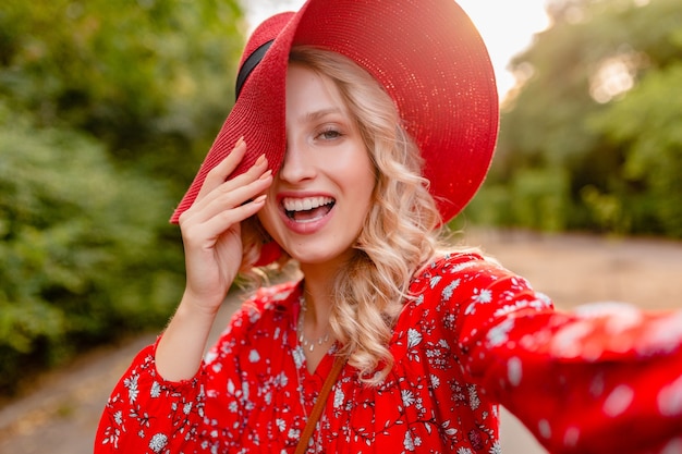 Mujer sonriente rubia elegante atractiva en traje de la manera del verano del sombrero rojo de la paja y de la blusa que toma la foto del selfie