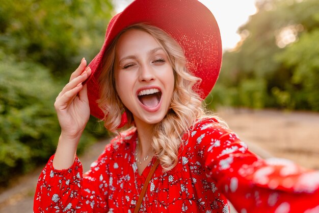 Mujer sonriente rubia elegante atractiva en traje de la manera del verano del sombrero rojo de la paja y de la blusa que toma la foto del selfie